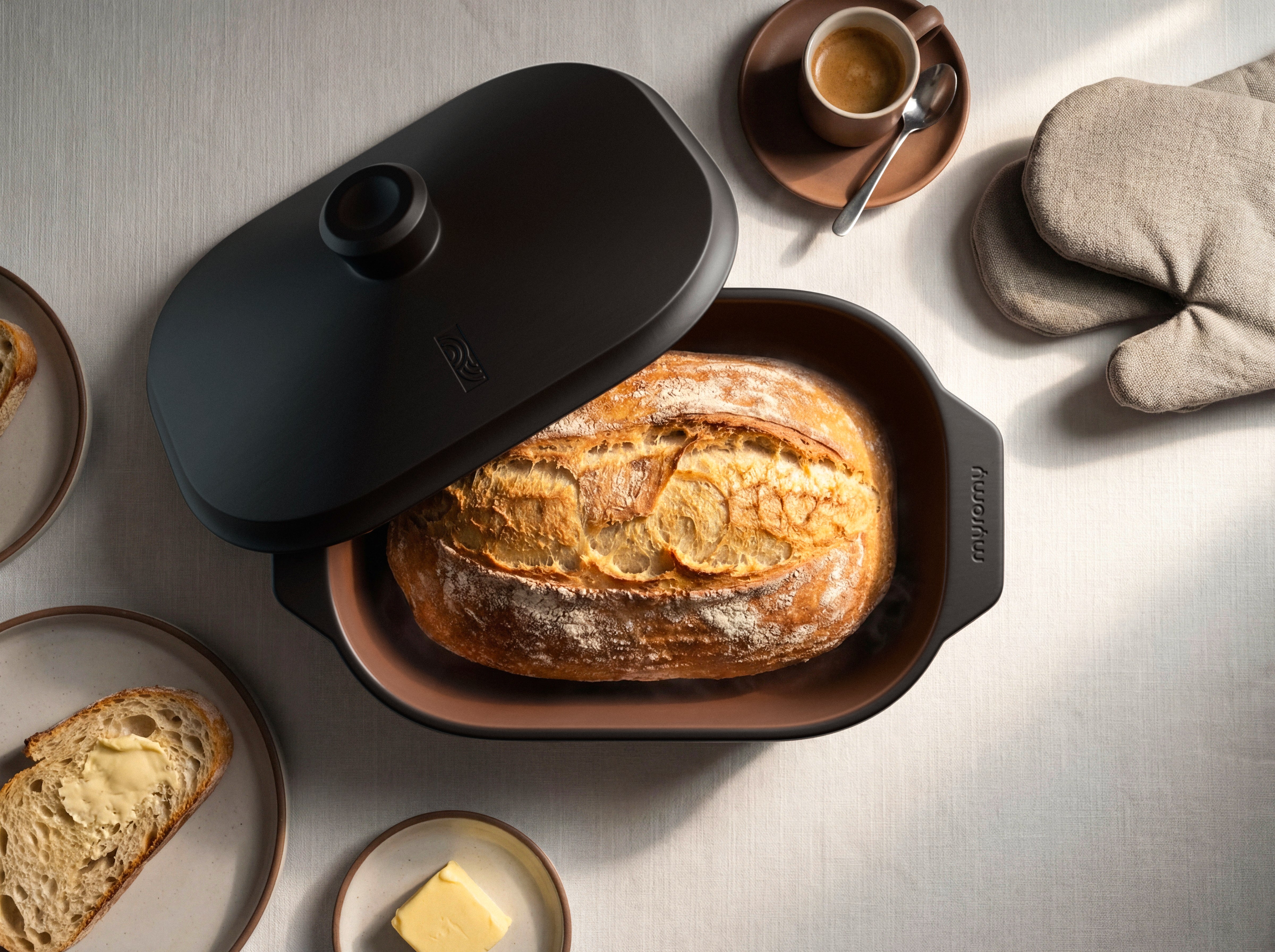 Frisch gebackenes Brot im geöffneten schwarzen myromy Multi-Bräter mit angelehntem Deckel, daneben Kaffee, Butter und Brotscheiben
