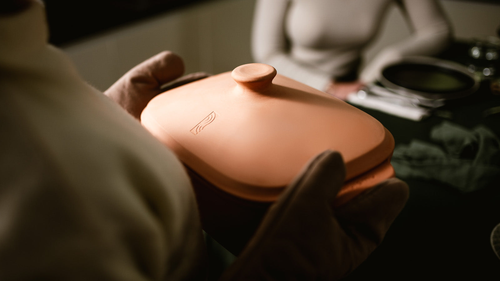 Person trägt den geschlossenen myromy Multi-Bräter in der Signature Edition als Keramik-Brotbackform und Keramik-Topf mit Ofenhandschuhen zum gedeckten Tisch