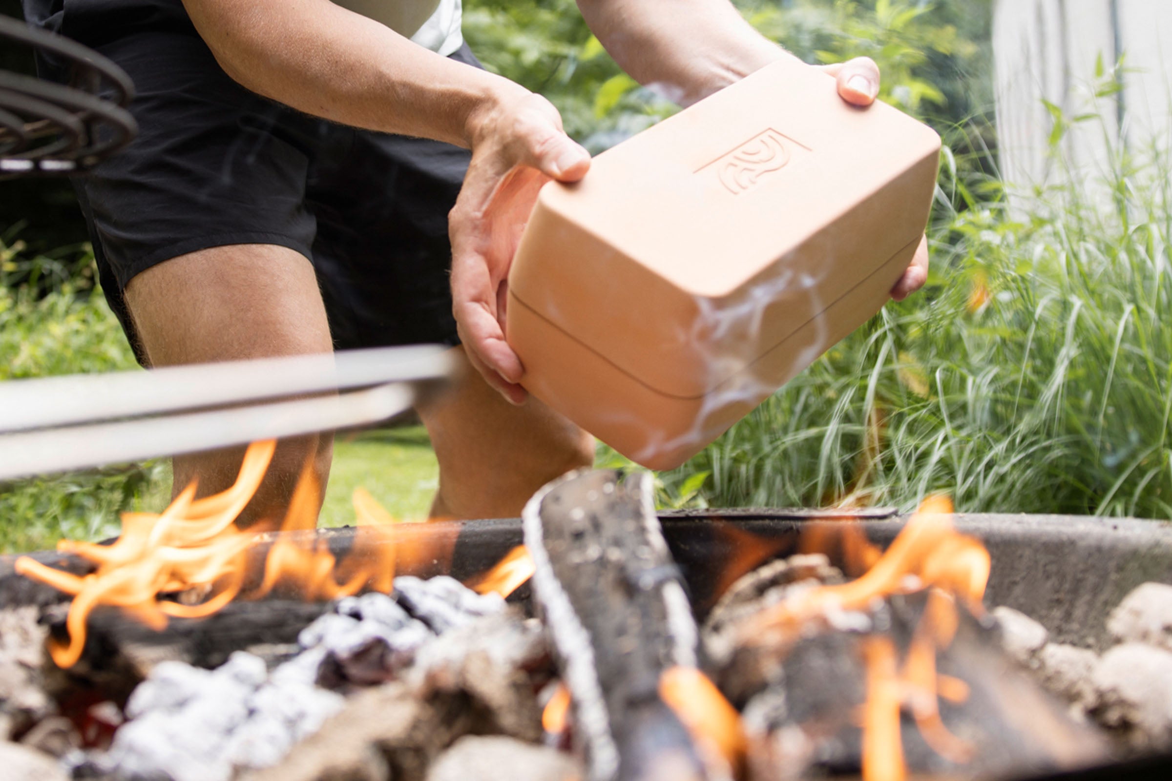 Person hält den geschlossenen myromy Brick BBQ Backstein aus Keramik über ein offenes Feuer im Garten