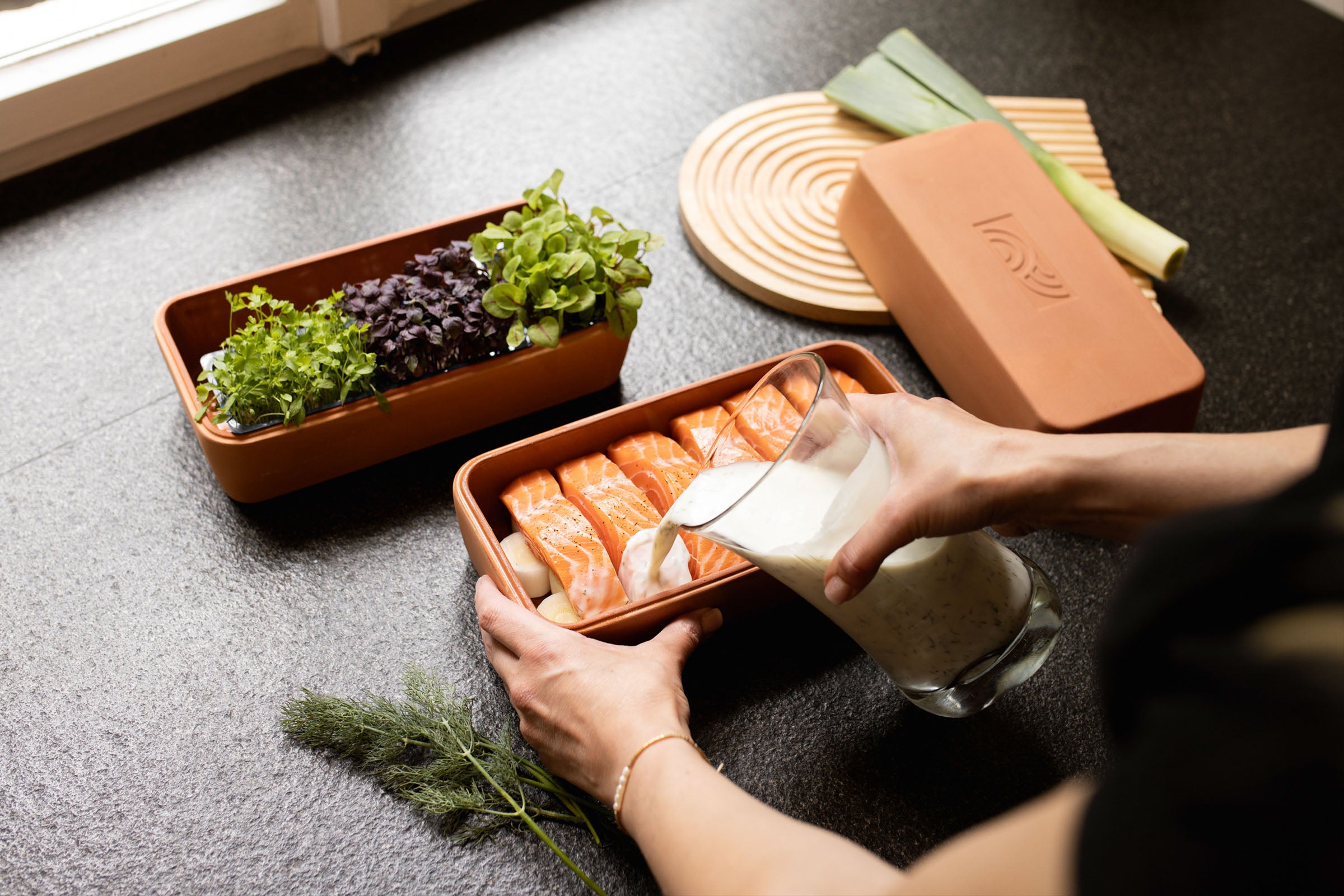 Sahnesauce wird über Lachsfilets im myromy Brick Classic Backstein aus Keramik gegossen, daneben stehen frische Kräuter und der Deckel
