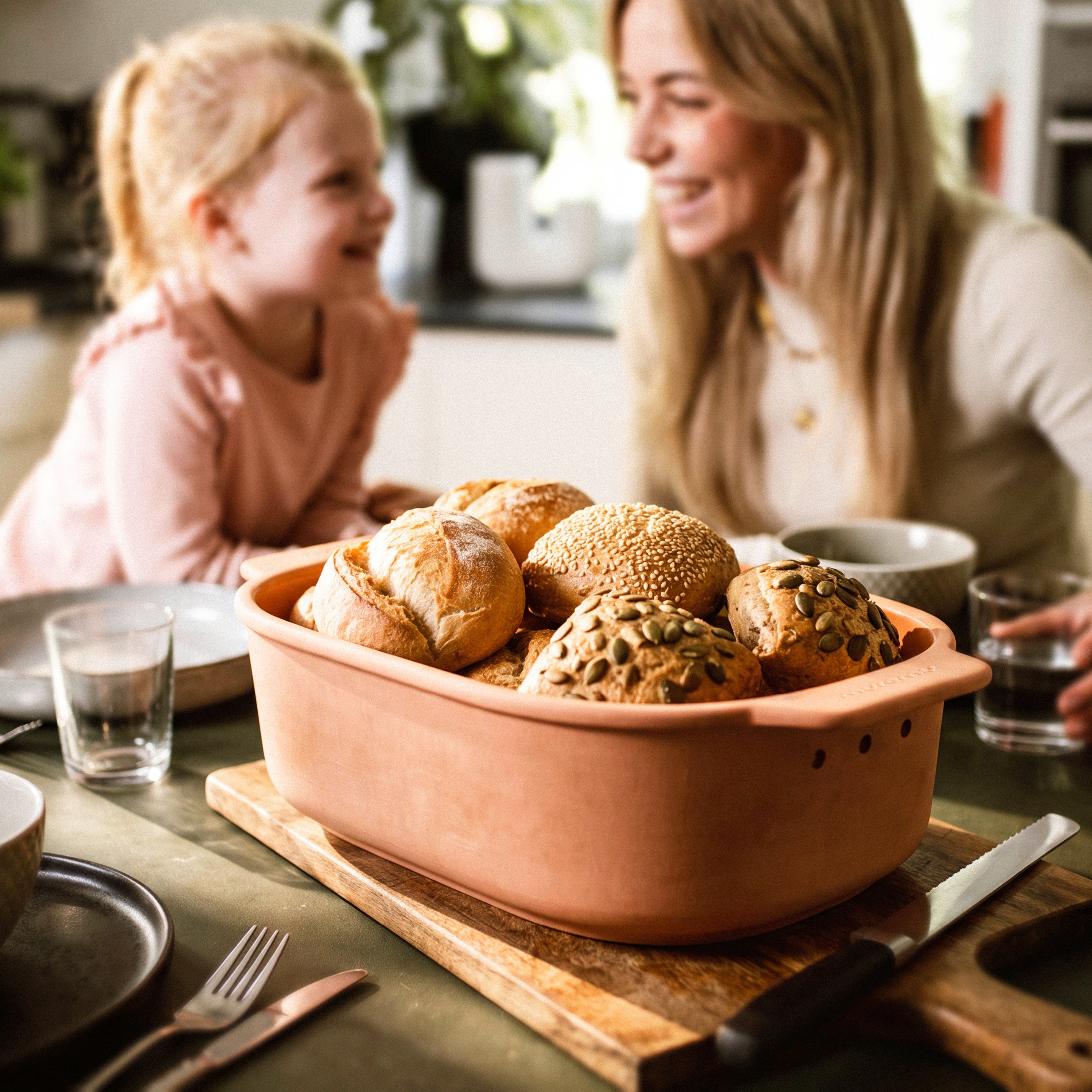 Brot-Frische-Topf Medium mit verschiedenen Brötchen auf einem Frühstückstisch, im Hintergrund sitzen Mutter und Kind