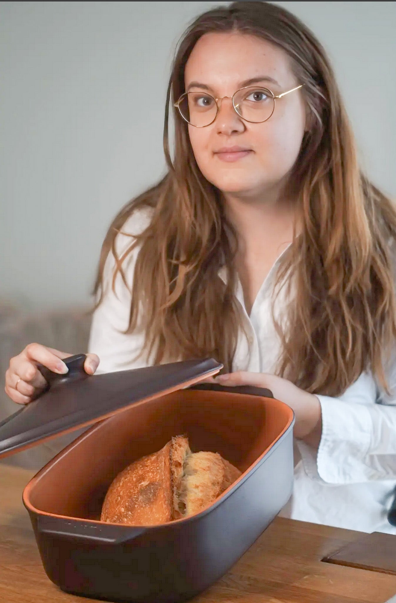 Frau öffnet einen schwarzen myromy Keramik-Brottopf und Brotkasten mit einem angeschnittenen Brot auf dem Tisch