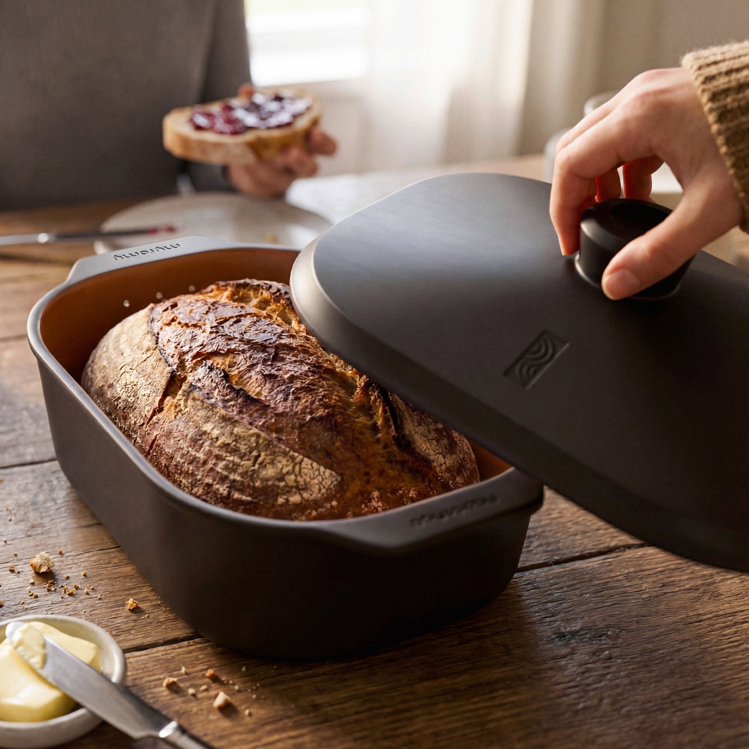 Eine Hand hebt den Deckel des schwarzen myromy Multi-Bräters als Keramik-Brotbackform und Keramik-Topf über einem frisch gebackenen Brot an