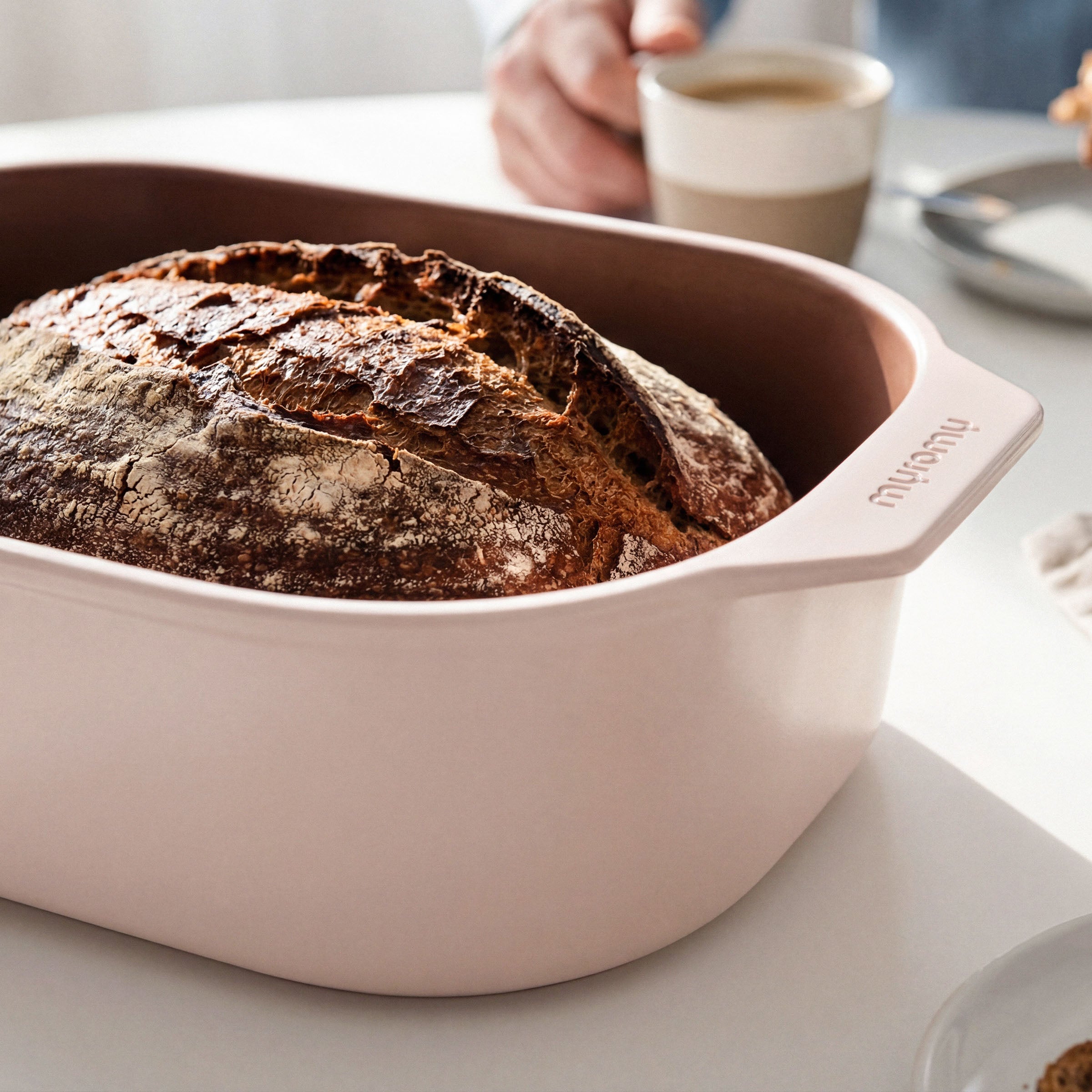 Frisch gebackenes Sauerteigbrot in einem weißen myromy Multi-Bräter als Keramik-Brotbackform und Keramik-Topf, daneben eine Tasse Kaffee